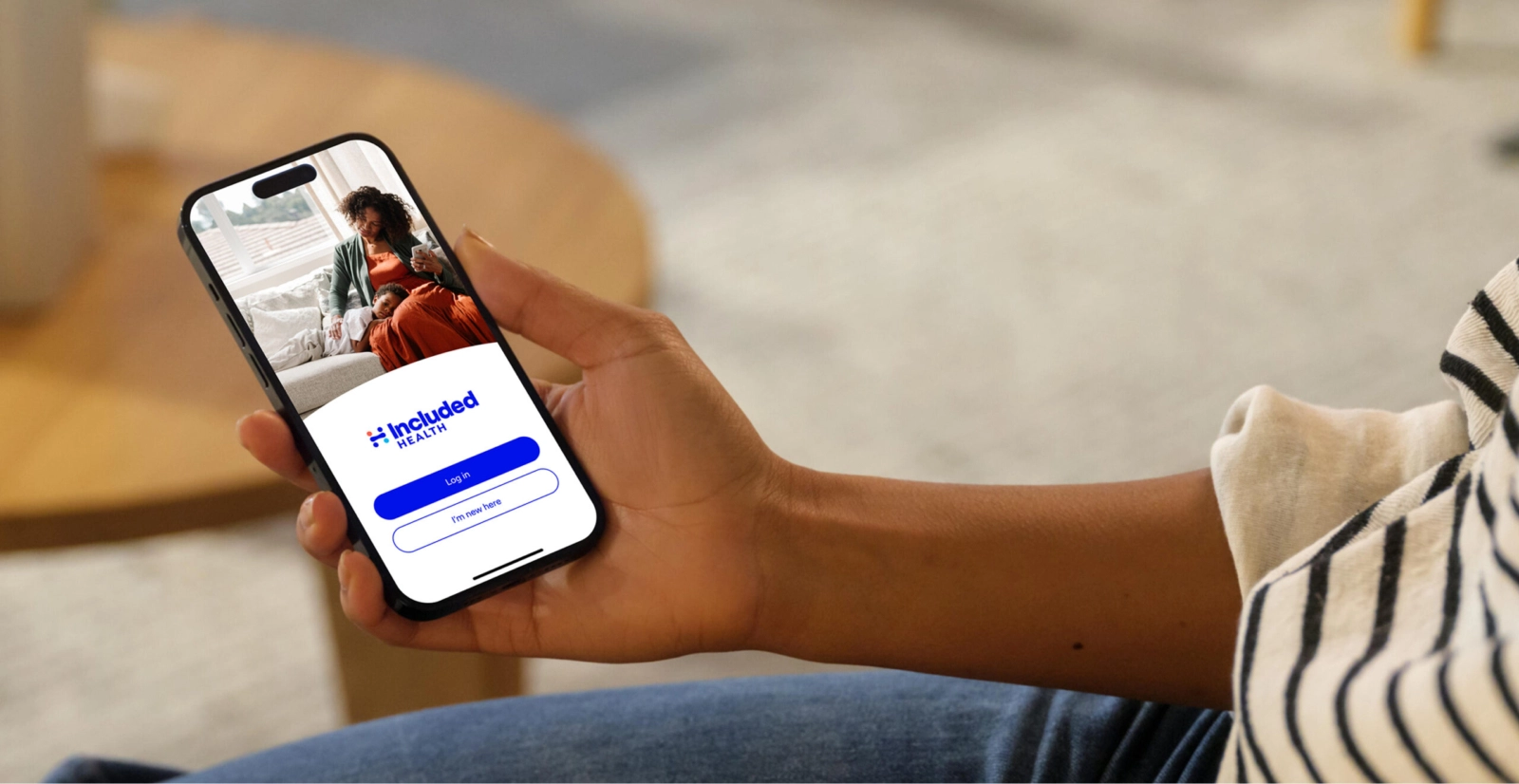 A close-up, angled view of a hand holding a smartphone. The screen displays the "Included Health" app's login page, with a video call between a mother and her child visible in the background.